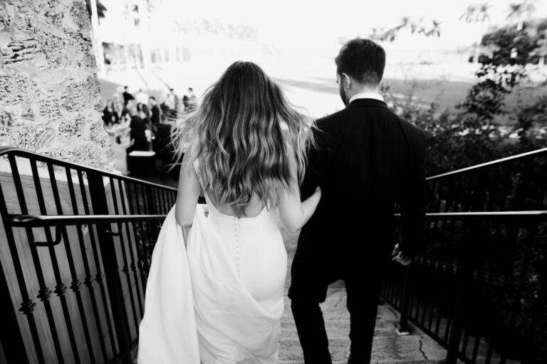 In black in white, a bride and groom walk down a staircase facing away from the camera.