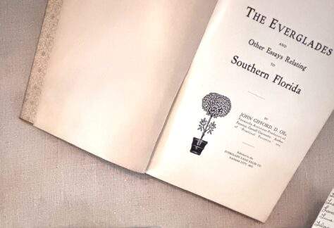 An open book rests on a light, textured fabric surface. The left page is blank, while the right page shows a title page with printed text reading “The Everglades and Other Essays Relating to Southern Florida.” Below the title is a small black illustration of a potted, round-canopied tree. Additional text on the page provides the author’s name and publishing information in smaller print. In the lower-right corner of the image, a separate small piece of paper with handwritten notes is partially visible.