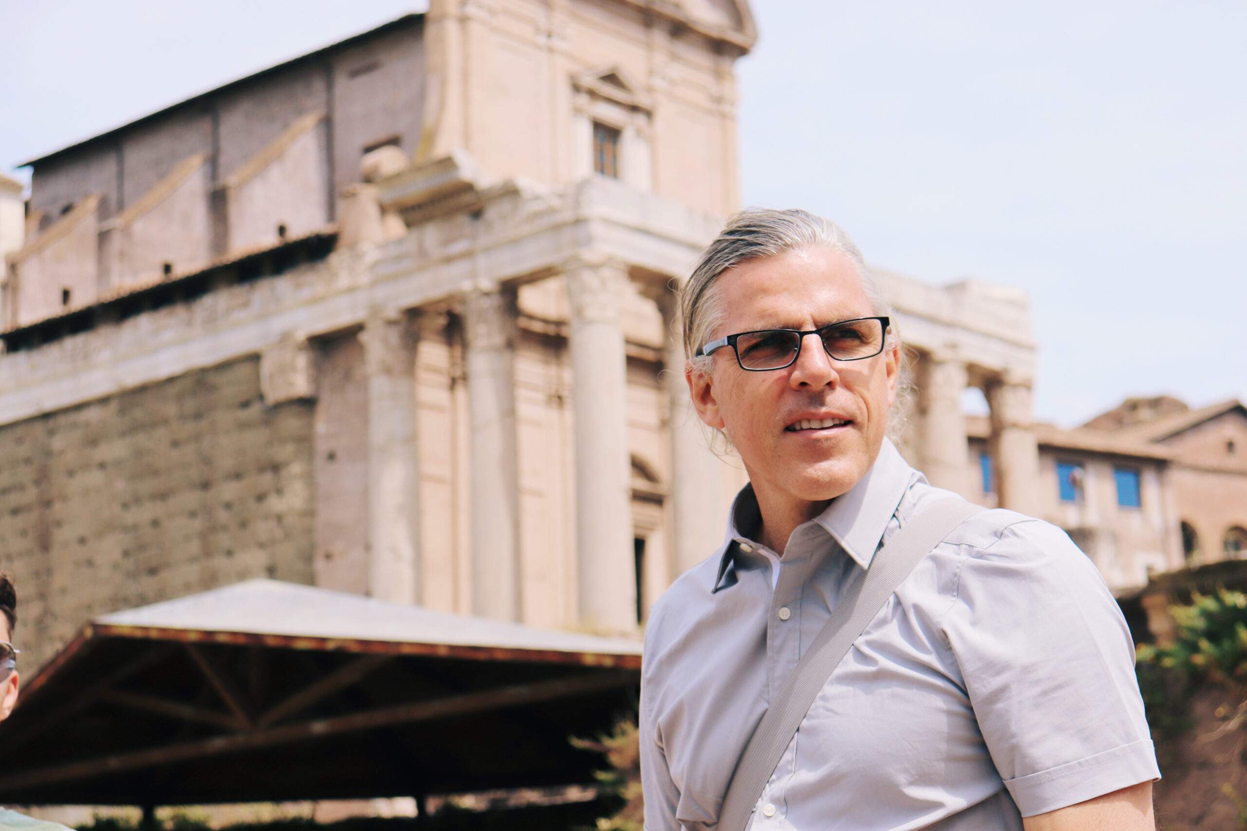 Historian John Bailly standing in front of the historic Roman Forum. He is grey haired, wearing black glasses and a grey button down shirt, with a grey-strapped bag across his body.