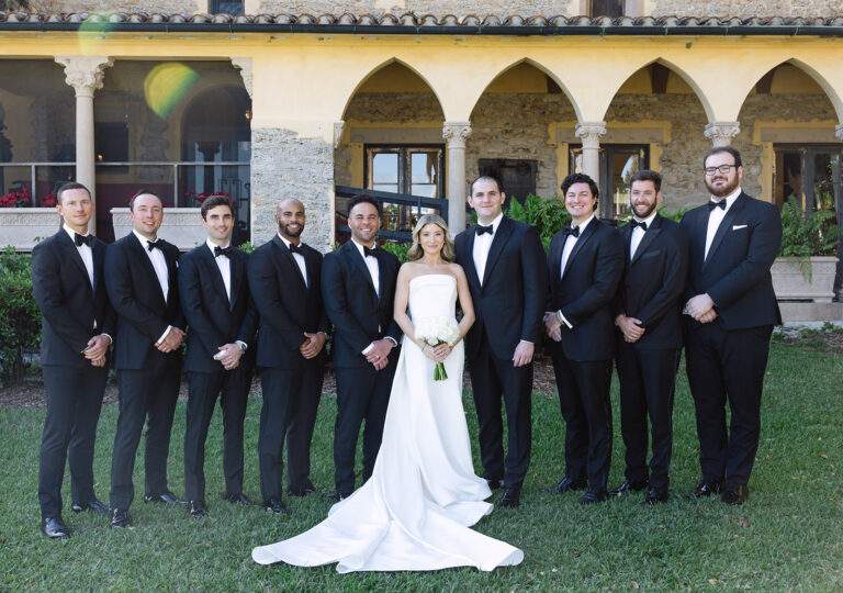 A bride in a long white dress holding a white boquet stands in the middle of nine groomsmen wearing black suits with bowties.